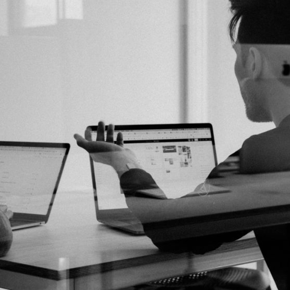 Schwarz-Weiß-Foto eines Entwicklers an zwei Laptops, Hand zeigt auf den Bildschirm