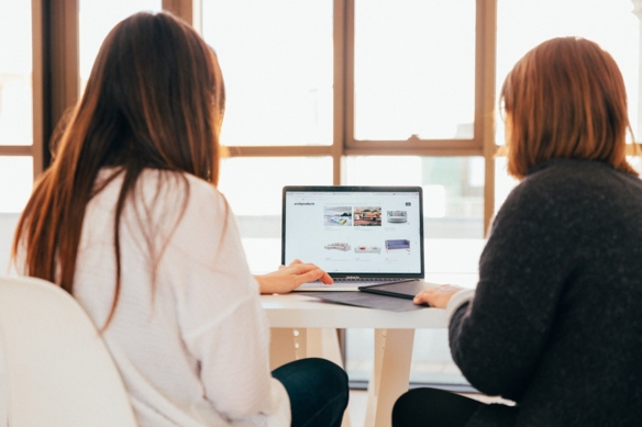 Zwei Frauen sitzen an einem Tisch mit einem Laptop und diskutieren.