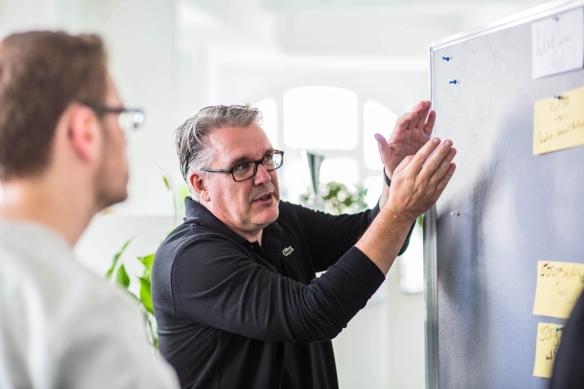 Logic Joe Managing Director erklärt etwas an einer Pinnwand mit Post-its, ein Kollege hört zu