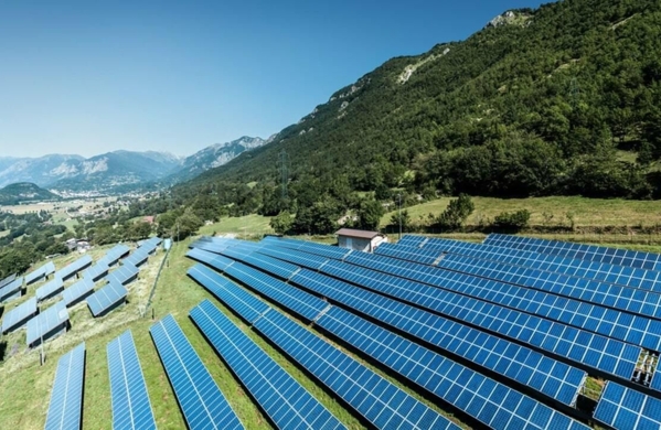 Luftaufnahme einer großen Solaranlage mit blauen Photovoltaikmodulen auf einem Berghang vor grüner Berglandschaft und blauem Himmel