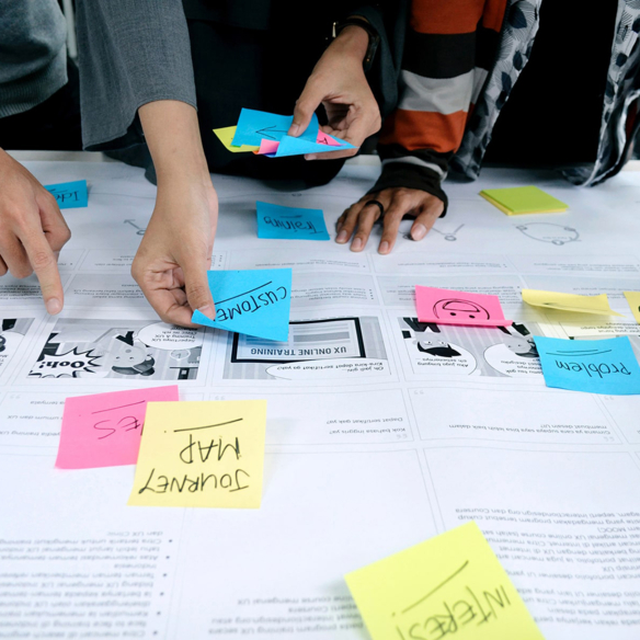 Hände von drei Personen arrangieren bunte Haftzettel mit Begriffen wie Customer, Journey Map und Interest auf einem Workshop-Tisch
