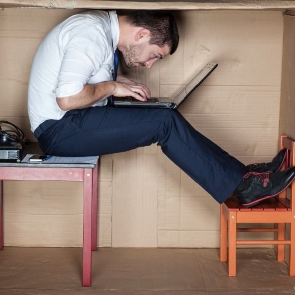 Ein Mann in Businesskleidung arbeitet an einem Laptop, eingepfercht in einem kleinen Pappkarton, die Knie an die Brust gezogen, zwischen einem Tisch und einem Stuhl sitzend.