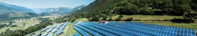 Luftaufnahme einer großen Solaranlage mit blauen Photovoltaikmodulen auf einem Berghang vor grüner Berglandschaft und blauem Himmel