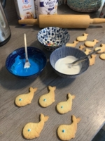 Selbst gebackene Walplätzchen beim Verzieren mit blauer und weißer Glasur in Schüsseln, Nudelholz und Puderzucker im Hintergrund